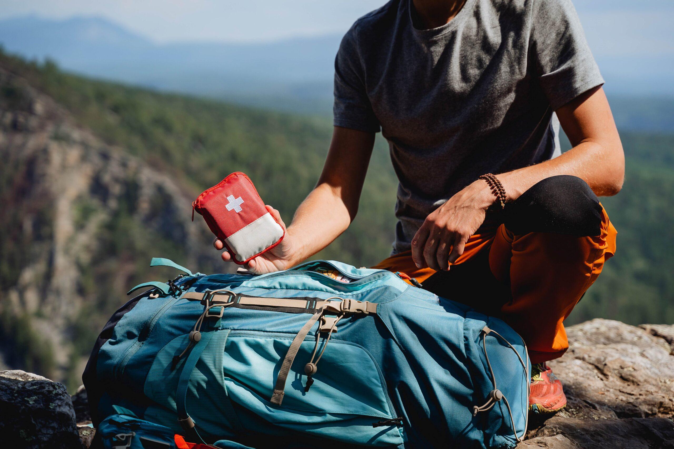 Ein Wanderer hält eine kleine, rote Erste-Hilfe-Tasche über einem gepackten Rucksack in einer malerischen Berglandschaft