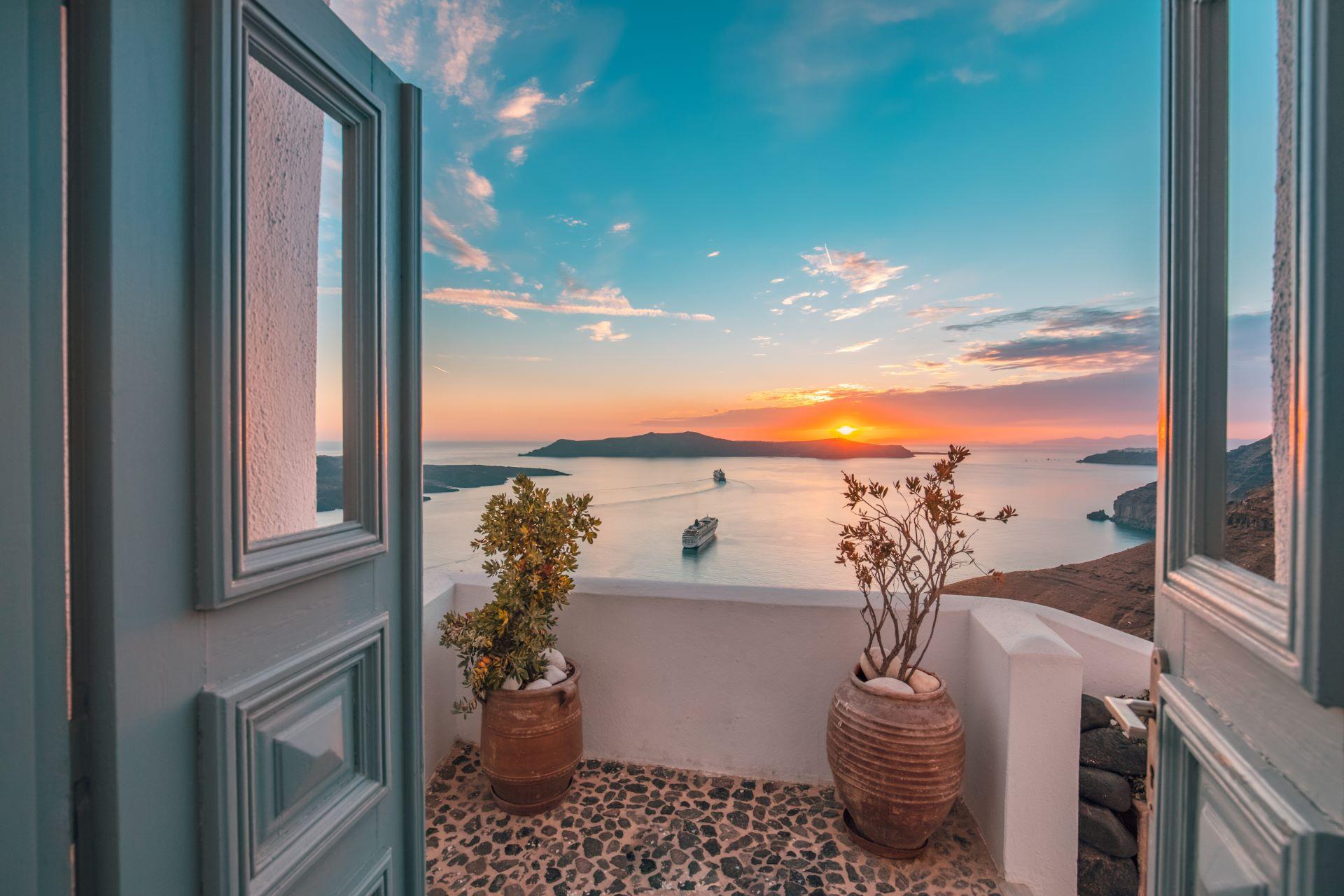 Blick von einer Terrasse in Griechenland auf einen atemberaubenden Sonnenuntergang über der Ägäis, mit einem Kreuzfahrtschiff auf dem Meer und zwei Pflanzen in Tontöpfen im Vordergrund.